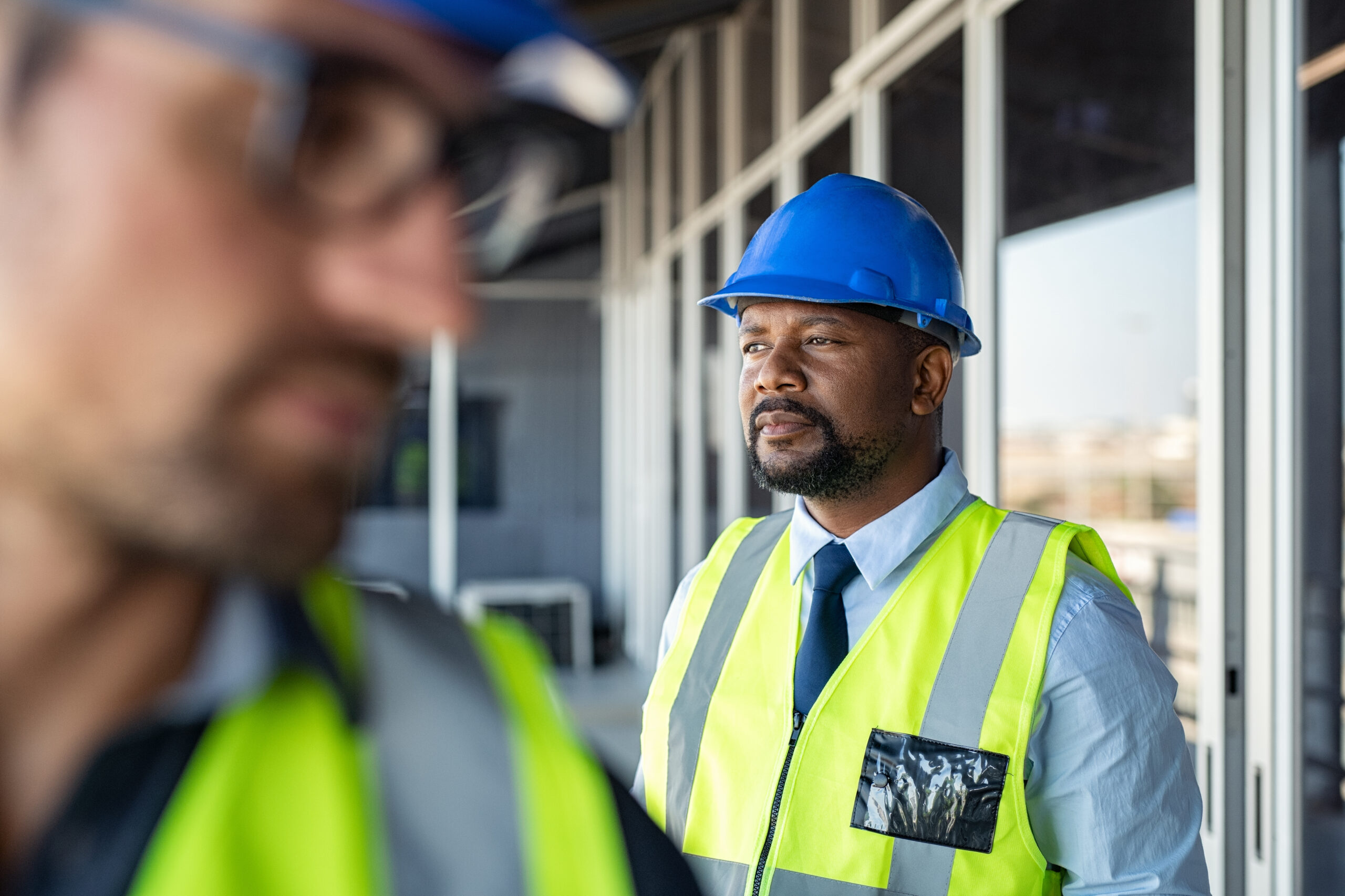 An African American man with a yellow vest and blue hard hat, looking out over a construction project